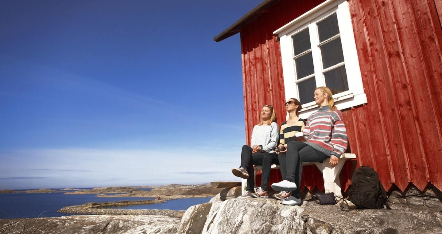 Three women sit on a bench outside of a red house. A seawall is seen in the distance.