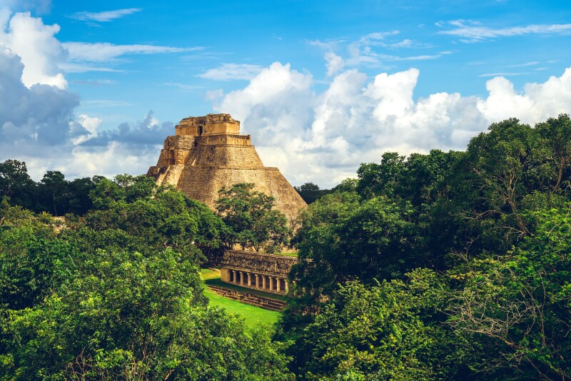 A large stone pyramid, with a smaller stone covered walkway in the foreground, surrounded by trees