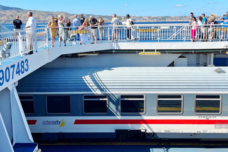 A blue, white, and red commuter train is being loaded onto a ferry to cross the Strait of Messina between Italy and Sicily. Passengers stand on the ferry deck above the train.