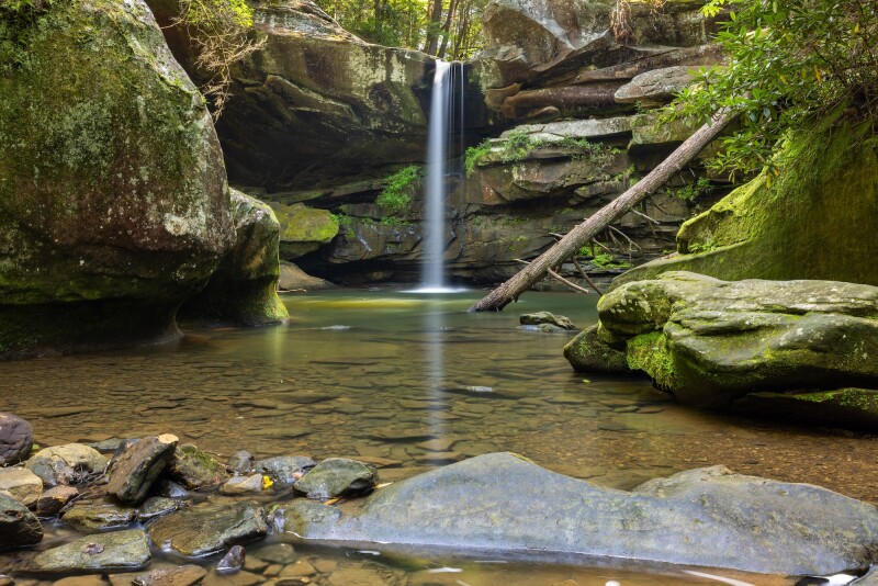 A long-exposure photograph of a tall, thin waterfall into a wading pool with moss-covered boulders and logs surrounding it