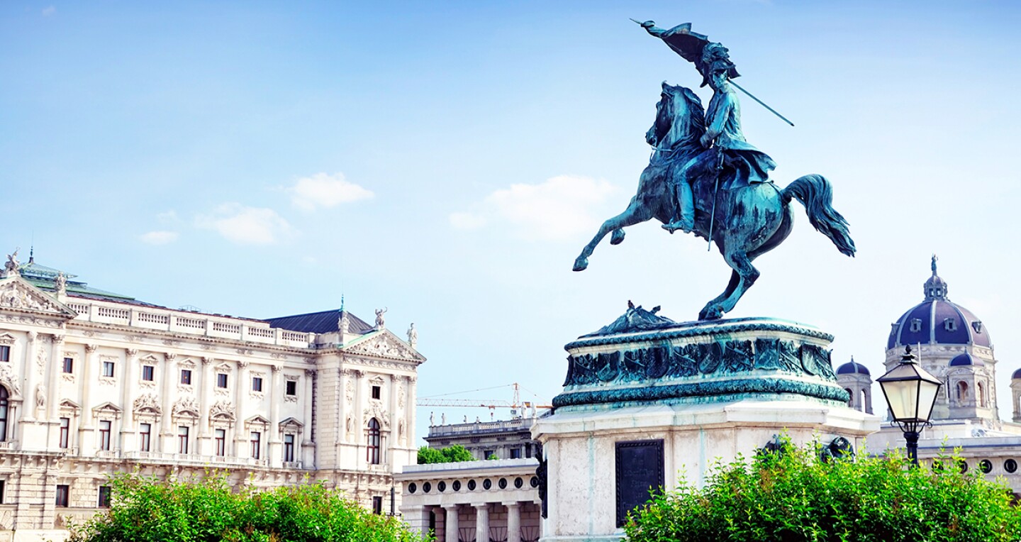A green copper statue of a man riding a horse. A large marble building is seen the backgrounds