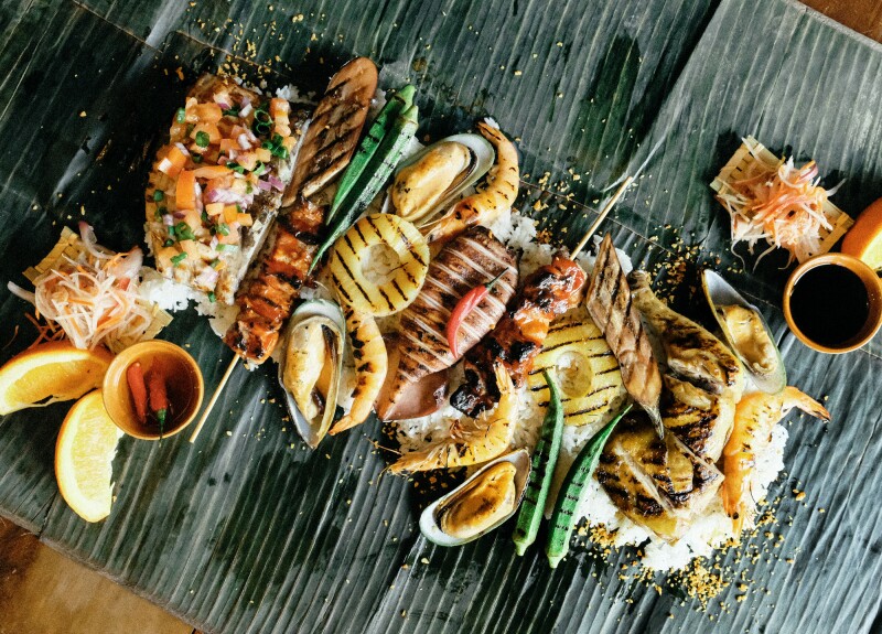 A mix of shellfish, fish, pork, vegetables, rice, and sauces spread out on banana leaves