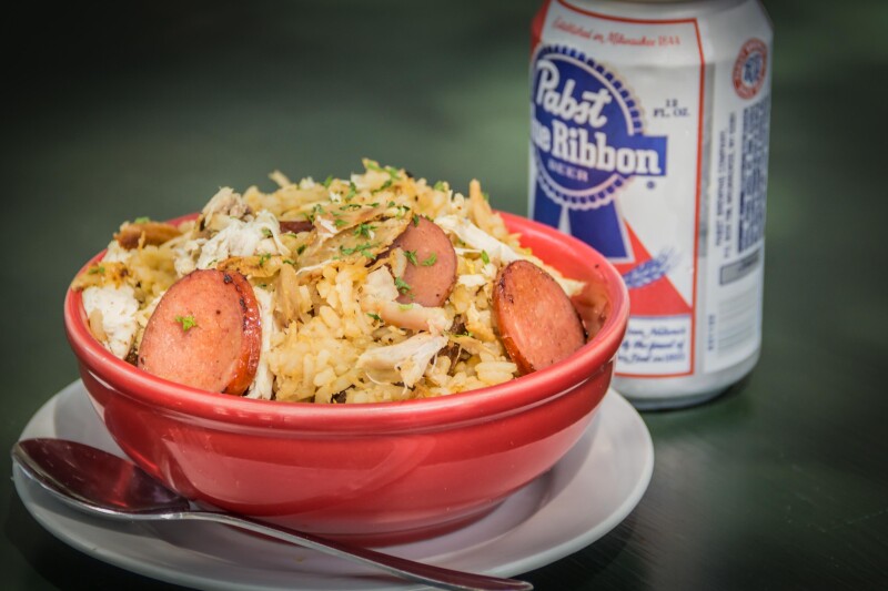 Can of beer beside a red bowl of rice, chicken, and sausage