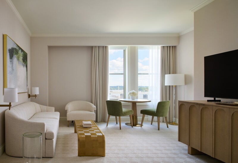 An elegant hotel room at the Salamander DC with wooden TV stand, green chairs near a window, a white couch, and a checkerboard-patterned table