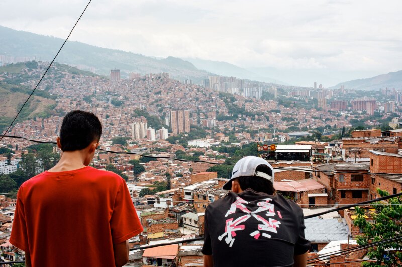 Two people wearing T-shirts seen from behind as they look out at a sprawling city of hills covered in brown and white buildings, "Tour Comuna 13", Comuna 13, Medellín