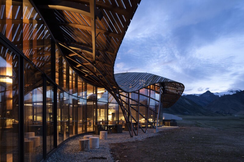 The Lindis Lodge has a large, curved roof and large glass windows. It lies in the Ahuriri valley. Large clouds cover the sky.