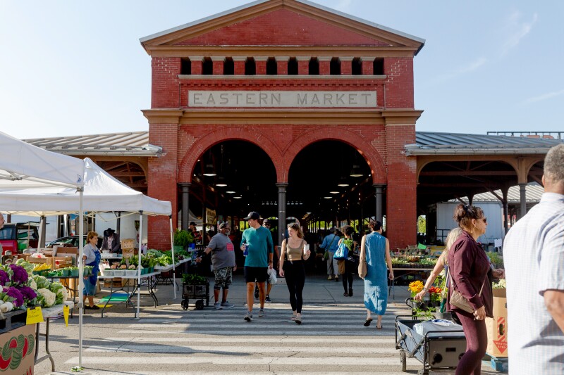 Red brick facade of Eastern Market, with double arches leading back to roofed area; shoppers and vendors outside in front of it