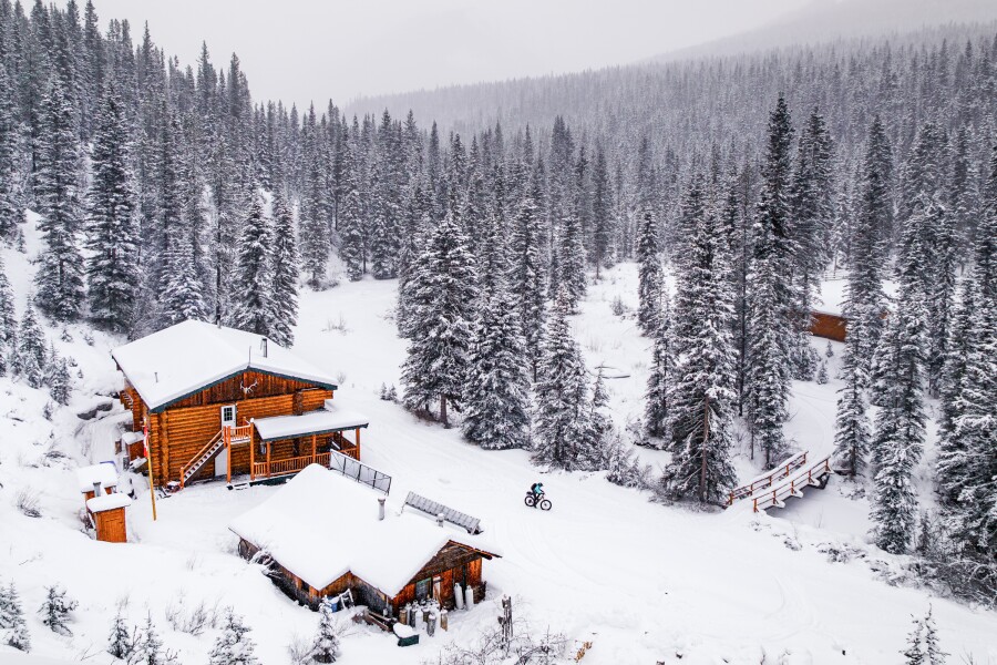 A person rides a bike over snow, past a wooden cabin in a snowy pine forest.