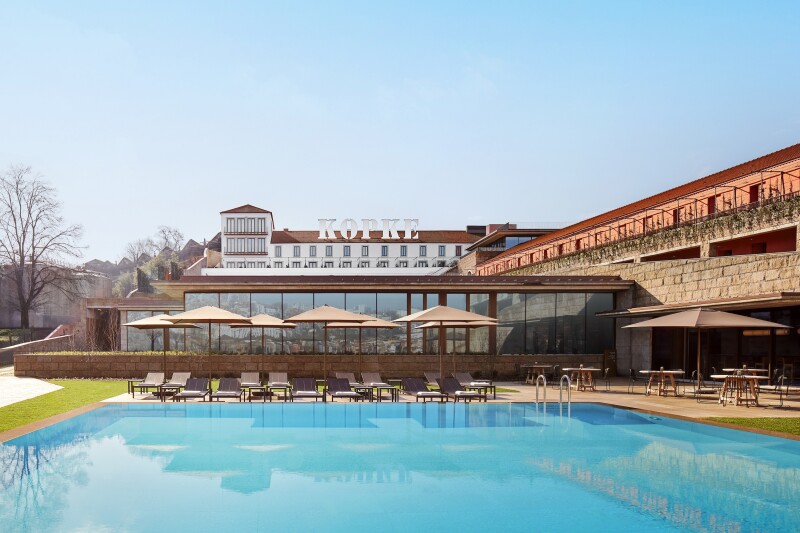 A pool area surrounded by lounge chairs and umbrellas, with the Tivoli Kopke Porto Gaia hotel building in the background and a large "Kopke" sign in white on top of it