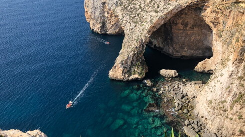 A single boat glides along the dark-blue water in Malta with a large, sand-colored cliff along the shore