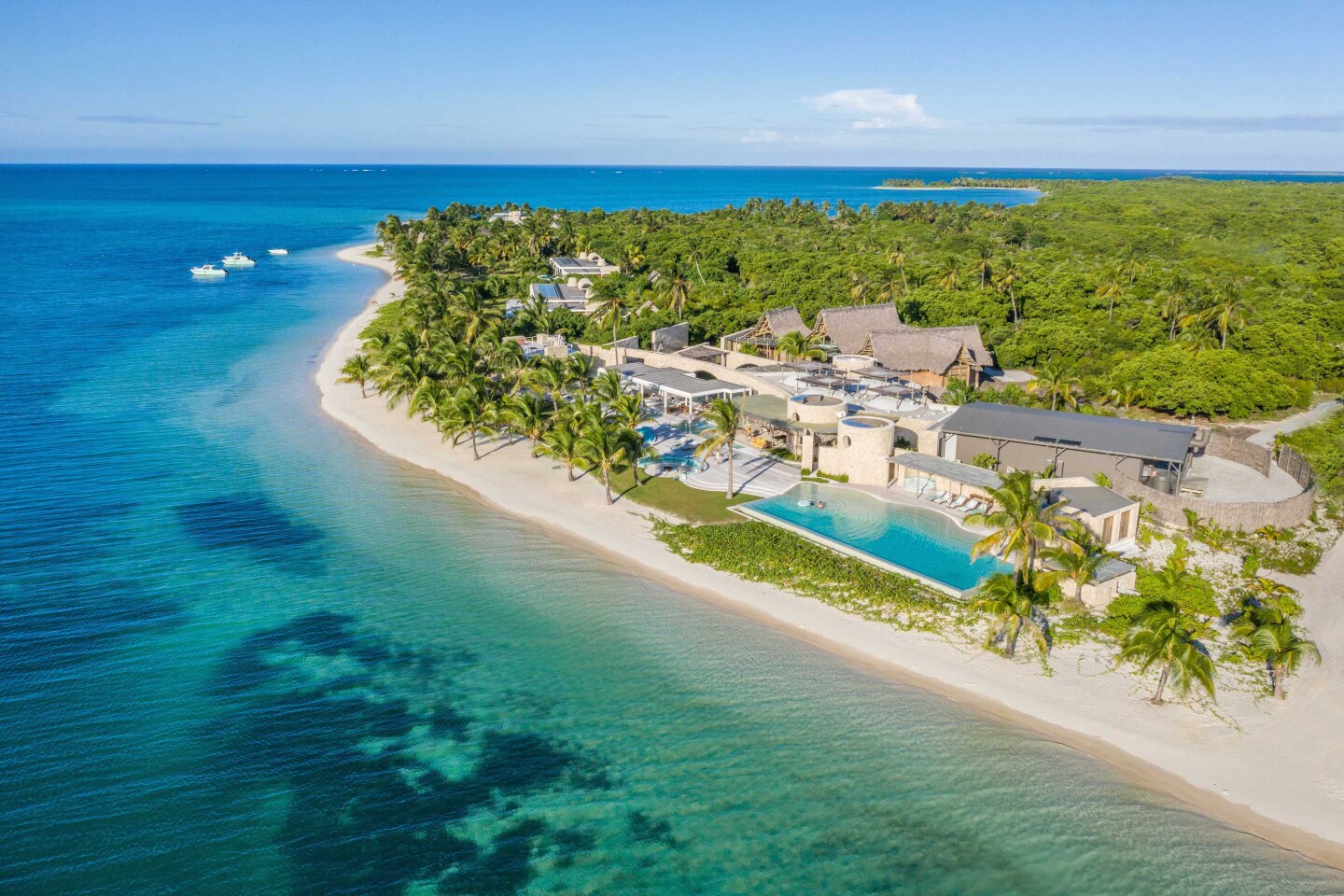 Aerial view of white-sand beach with clear seawater, palm trees, buildings, and long, rectangular pool