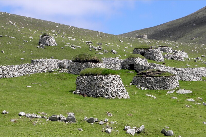 Distant view of stone walls and small round shelters (with grass roofs) in green field