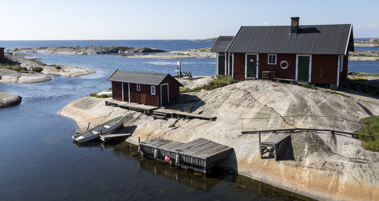 A house on a rock by the sea. A modest wooden boat rests against the shore by a shed.