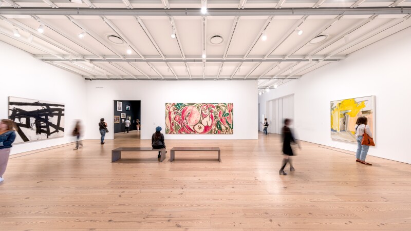 A wide gallery with white walls and several people viewing large modern paintings