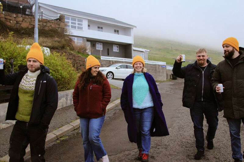 Five people in jackets and yellow beanies walking down a street in the Faroe Islands, with modern house in background