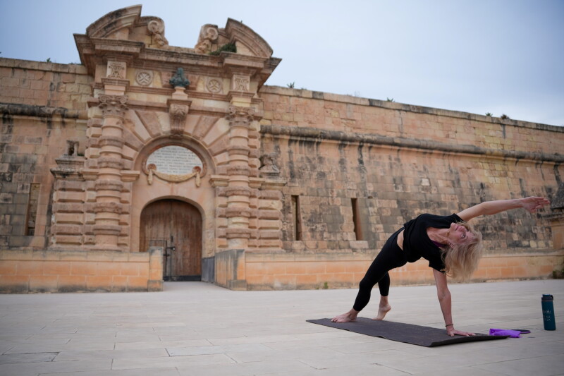 A blonde woman in black yoga clothes on a yoga mat in camatkarasana (wild thing pose) with a water bottle and towel at Fort Manoel