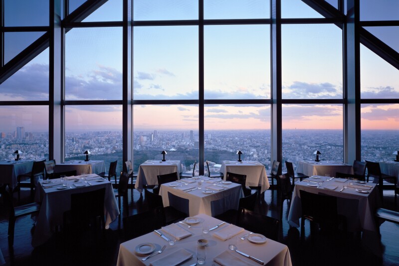 Empty tables set for dinner at the New York Grill at the Park Hyatt Tokyo with high ceilings and a wall of windows overlooking the Tokyo skyline, pictured at sunset