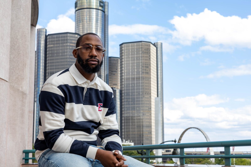 Photographer Elonte Davis sitting next to a railing in front of the Detroit skyline wearing a blue and white striped rugby shirt, blue jeans, and light brown sunglasses