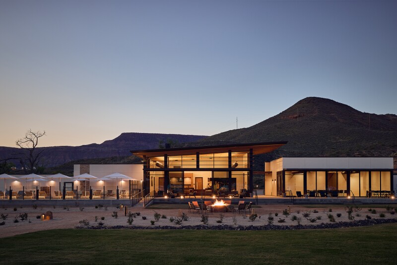 Outside of the AutoCamp Clubhouse, a mid-century modern building, at night time, with a firepit that has a fire in it in front.