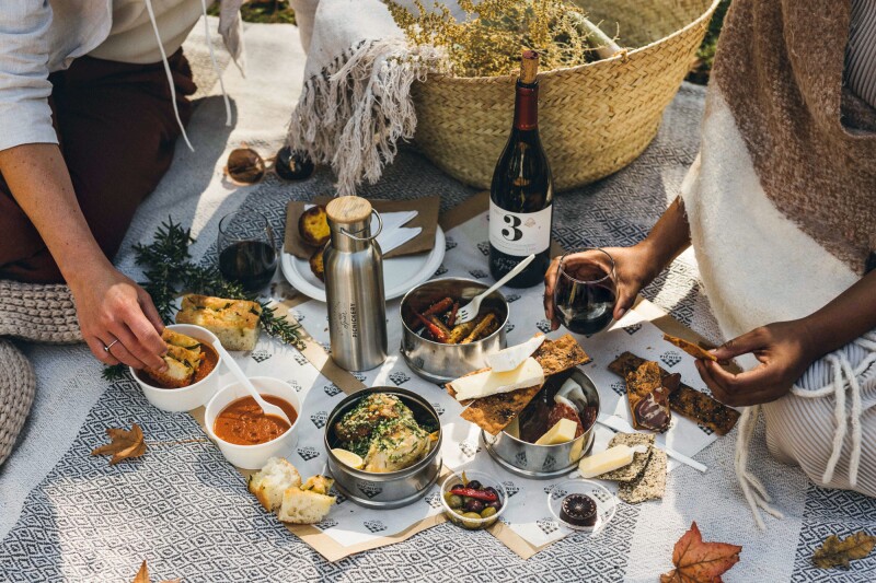 Two people, only their arms and hands in view, sit on a blanket enjoying a picnic with wine and containers of food.