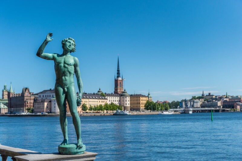 A green statue of a nude man in front of a body of water in Stockholm Harbor, Sweeden.