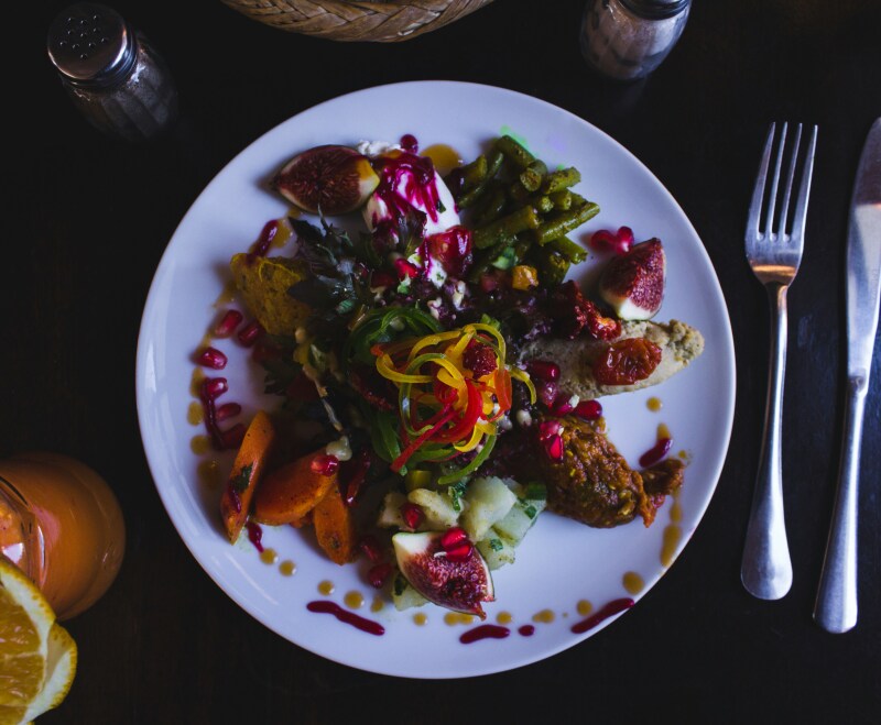 A white plate filled will colorful vegetables, figs, and pomegranate seeds, viewed from overhead, next to fork and knife