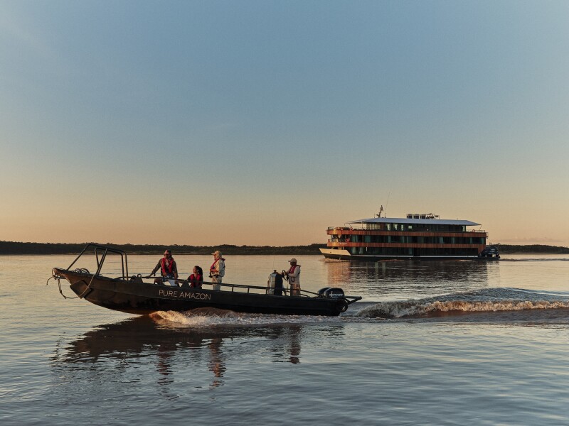 A skiff with four people on board rides in front of the 'Pure Amazon' riverboat, which is in the background, during sunset