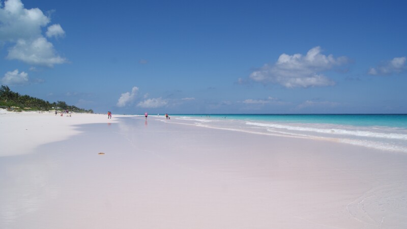 Beach with pale pink sand, blue skies, and a light blue water; a few people in distance