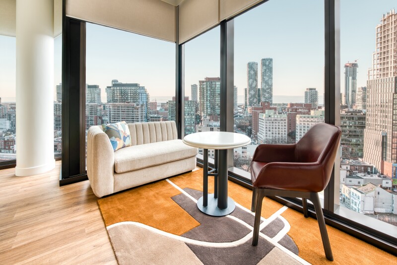 Corner guest room, with walls of windows overlooking city, pale wood floor with abstract patterned rug (orange, brown, tan), and modern furniture