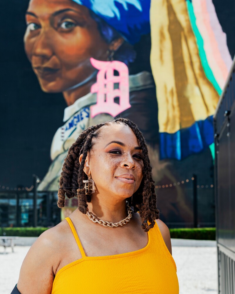 Sydney G. James, wearing yellow top, stands in front of her mural "Girl with a D Earring"