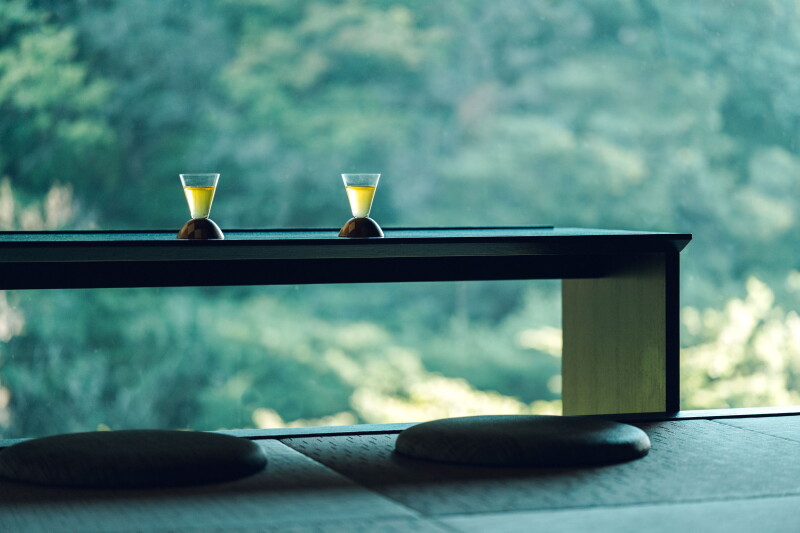 Two small cups filled with a yellowish beverage sit on a low table overlooking a forest.