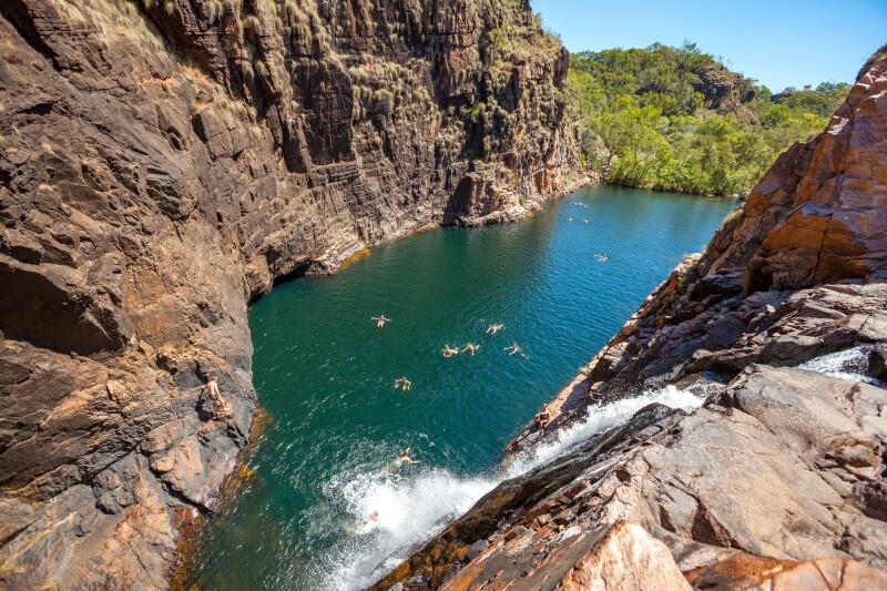 Distant view of several people swimming in a turquoise natural rock pool, with waterfall at right