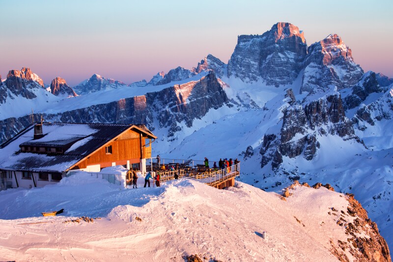 Distant aerial view at dusk of people on an observation deck surrounded by snowy, dramatic mountains