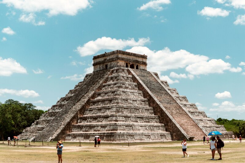 Stone square pyramid with steps up the middle of each side sits in a field as tourists wander around taking photos on a sunny day