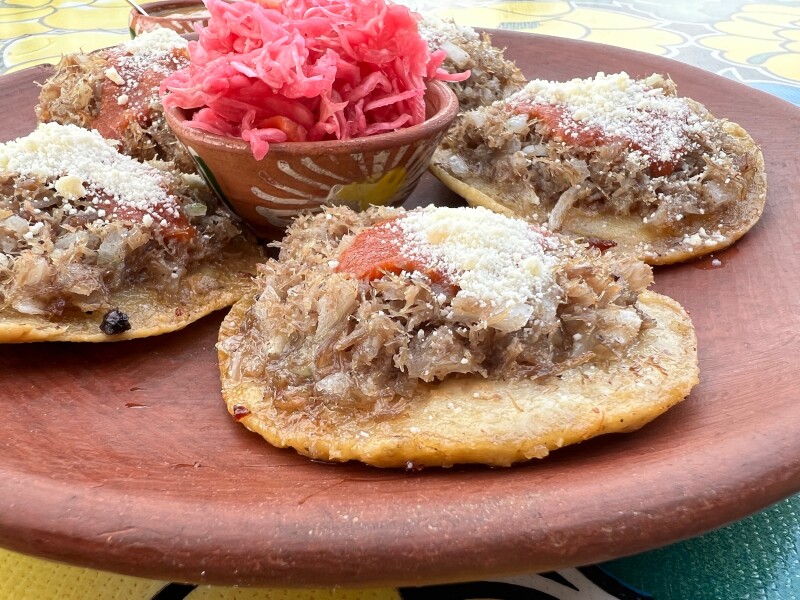 A platter of garnachas, fried corn tortillas topped with stewed shredded meat, queso fresco, and salsa, with bowl of red cabbage slaw in center