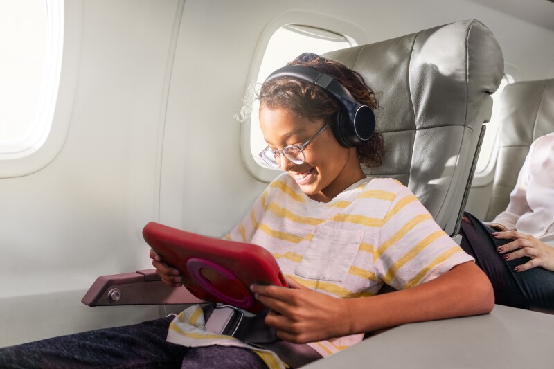 A child with iPad in red case sitting wearing headphones and eyeglasses and sitting in window seat on JSX airplane