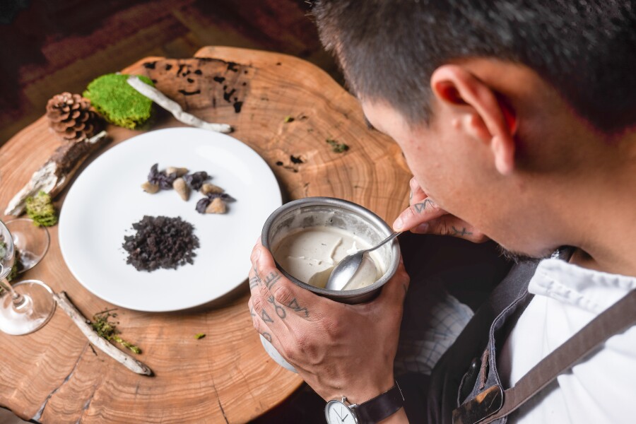 A chef holds small container in left hand to stir sauce for plate of food atop tree stump