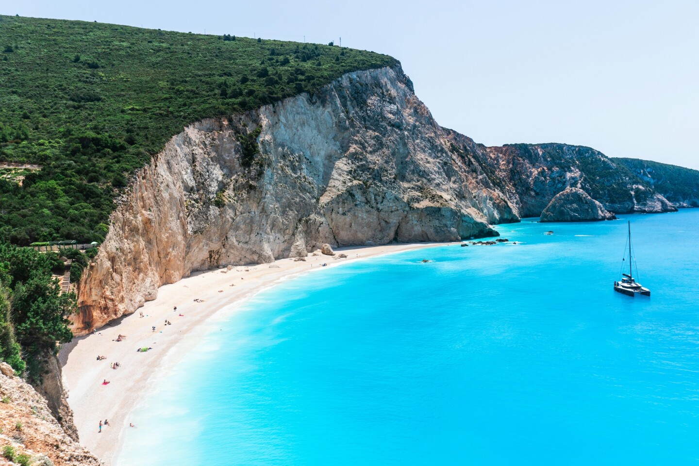 A catamaran in neon blue water off a white-sand beach with cliffs in the background