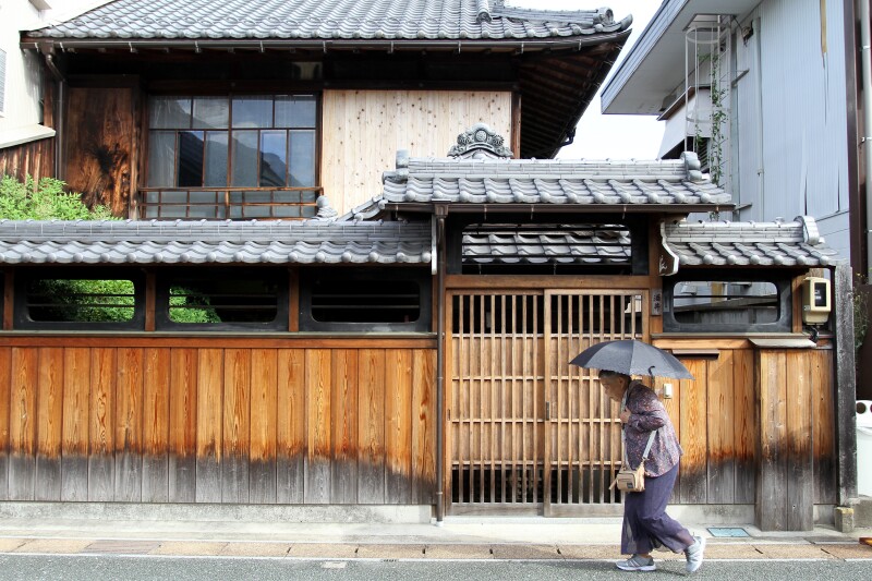 Exterior of old house in Udatsu, with wooden fence and gray tiled roofs; a person using black umbrella is in foreground