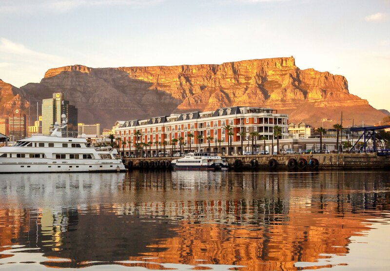 A brick hotel next to a marina, with boats and water in foreground against backdrop of Table Mountain on Cape Town waterfront