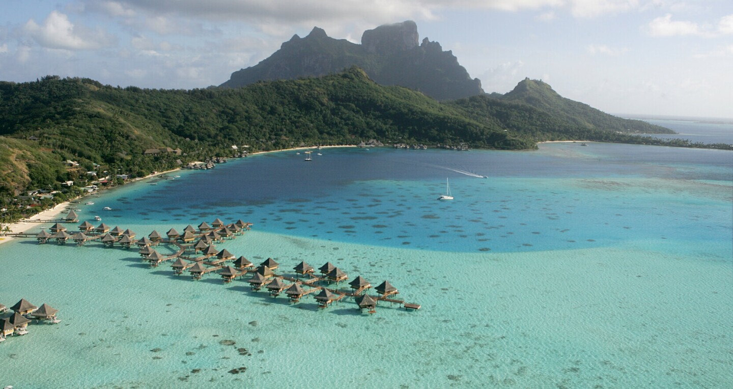 The private bungalows of the InterContinental Bora Bora Le Moana Resort sit above the expansive turquoise water, with a mountainous landscape in the background.
