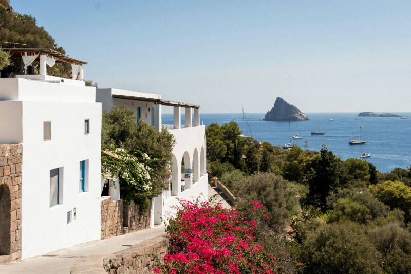 White-painted Mediterranean house, with red bougainvillea and green foliage at right, overlooking sea dotted with moored yachts and jagged, rocky island.