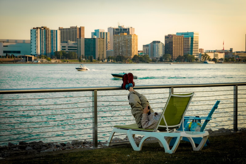 A person lounging in a green and white lawn chair with their feet up on a railing takes in the view of the Detroit River and skyline from Belle Isle Park