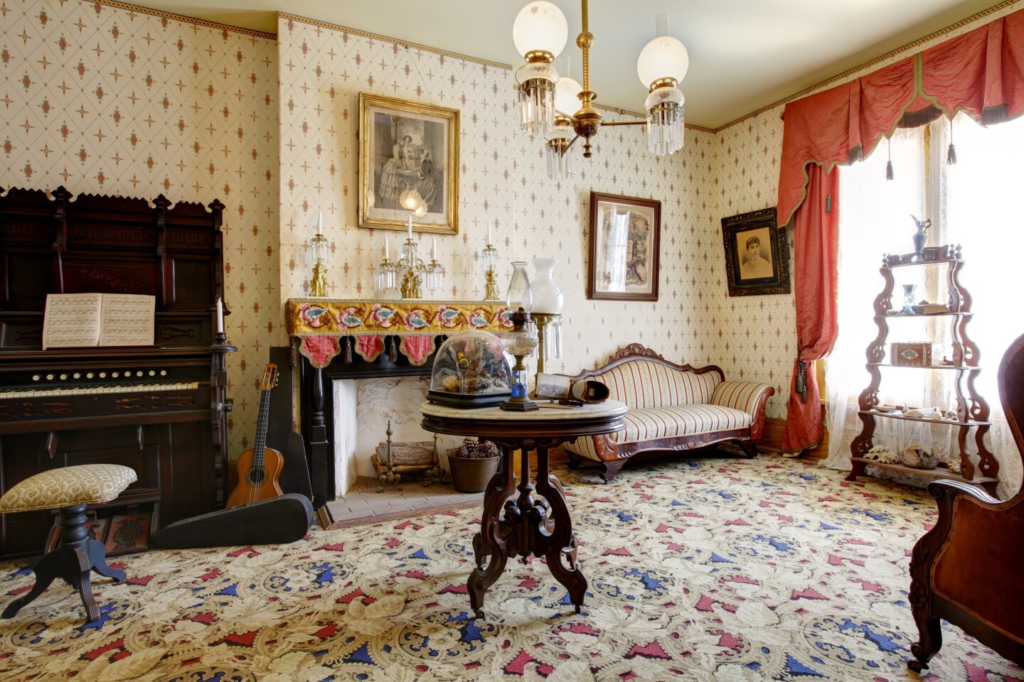 A room in an 1857 house with a patterned carpet, patterned wallpaper, a large wooden piano, and a black marble fireplace.