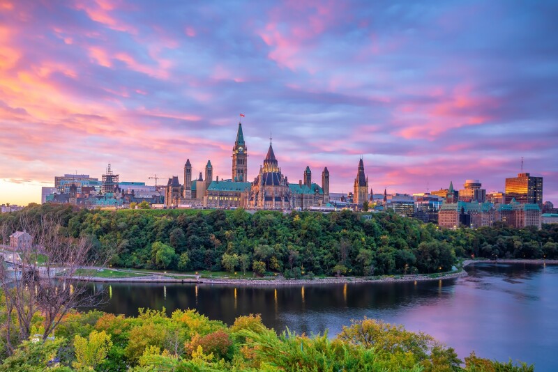 Parliament Hill in Ottawa at sunset, with river, roadway, and trees in foreground