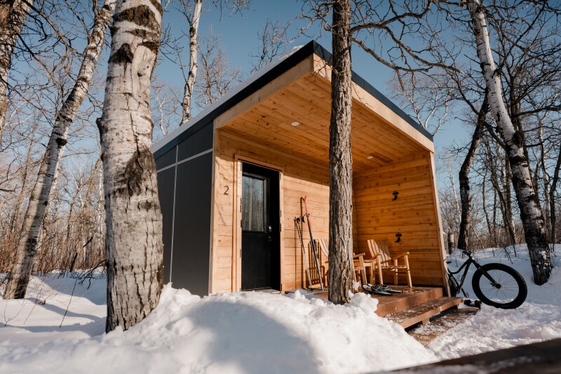 A bike leans against the side of a tiny wood cabin with a small covered front patio on which are two Adirondack chairs, surrounded by snowy forest
