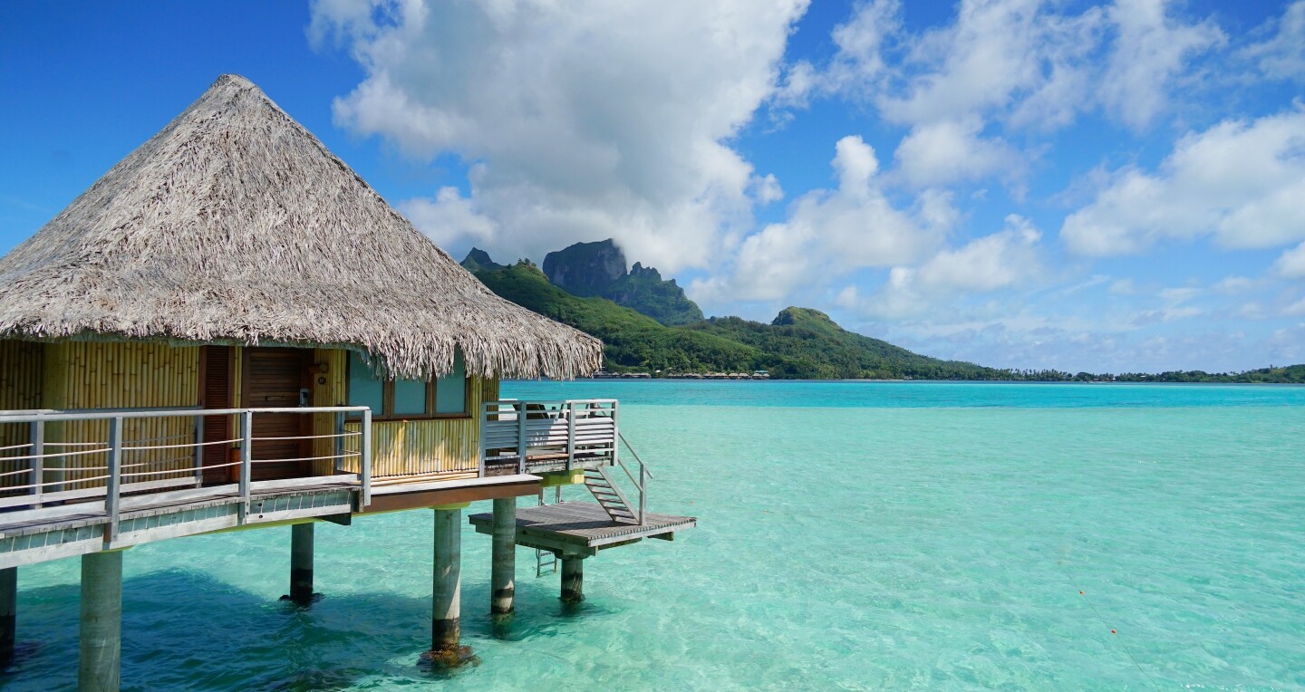 A single private bungalow sits above turquoise water. Stairs lead down to a swimming platform, and the mountainous landscape looms in the background. It is a sunny day and some clouds mix in with the blue skies.