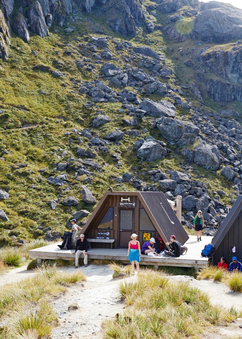 People lounging on the wooden patio of a flat-topped Harris Saddle warming hut in a mountainous setting
