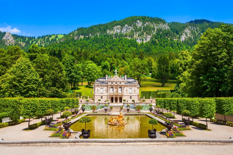 Linderhof Palace in southwest Bavaria, Germany, seen with soaring mountains in the background and a pond with a golden statue in the middle and a manicured garden all around