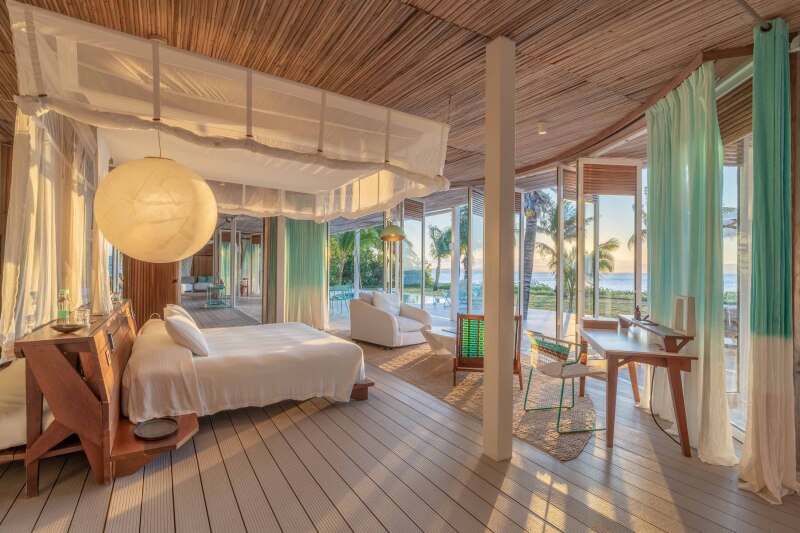 The ocean-facing master bedroom of a private villa at Miavana at Nosy Ankao, Madagascar, which includes a canopied bed, blue and white curtains, and several chairs for lounging.